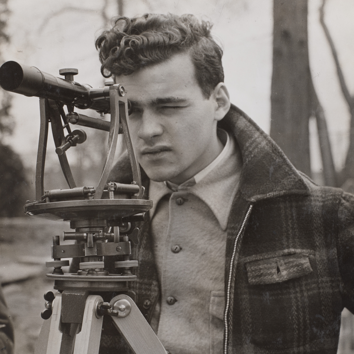 Young man in a plaid jacket looking through a surveying instrument on a tripod outdoors.