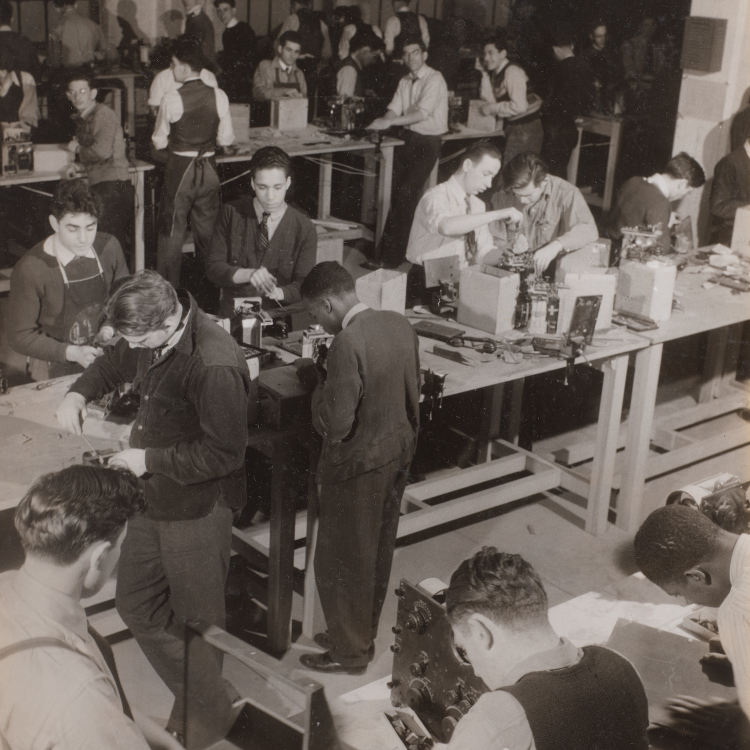 Black and white photo of men working on electronics at tables in a factory or workshop.