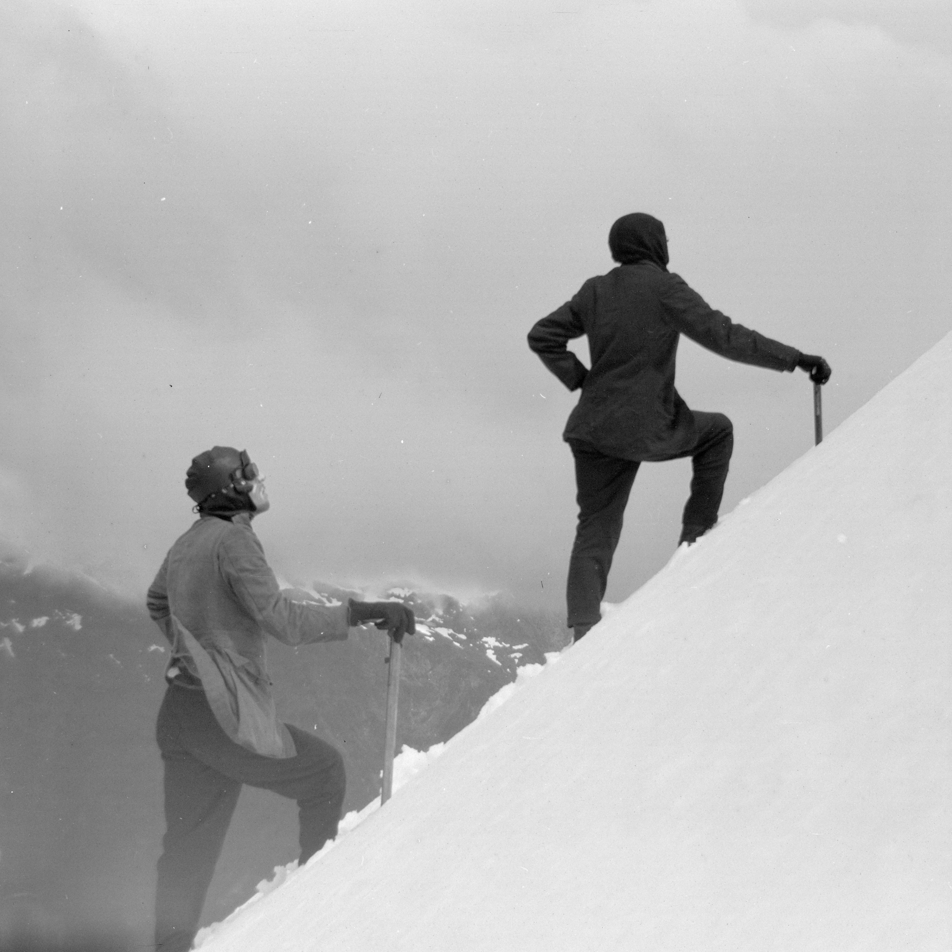 Two climbers ascending a steep snowy slope with mountains and cloudy sky in the background.