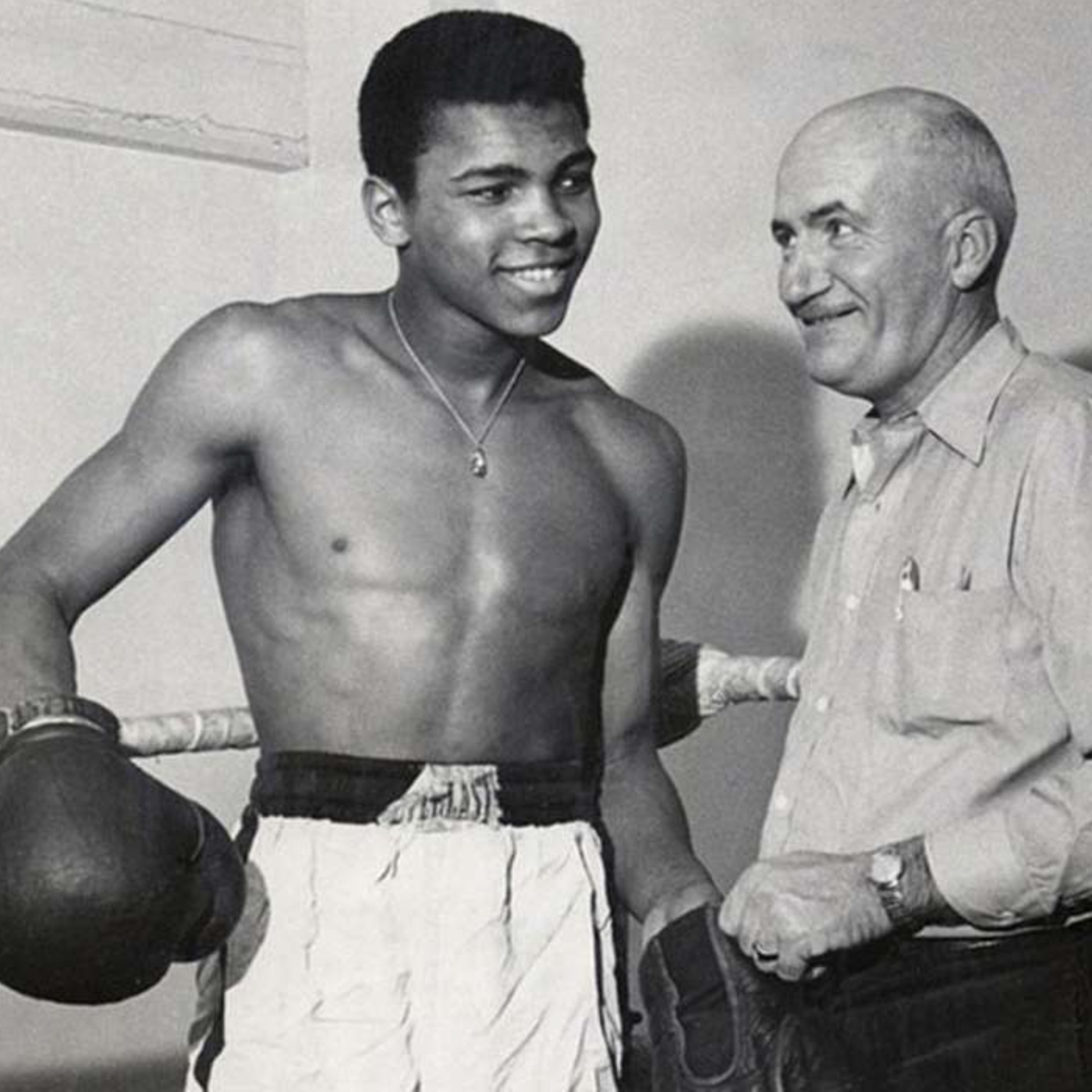 Young boxer smiling shirtless with gloves, standing next to his older coach in a gym.