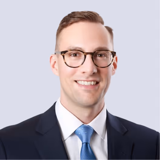 Smiling man wearing glasses, a dark suit, white shirt, and blue tie against a light background.