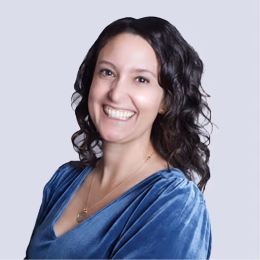 Smiling woman with curly dark hair wearing a blue velvet top against a light background.