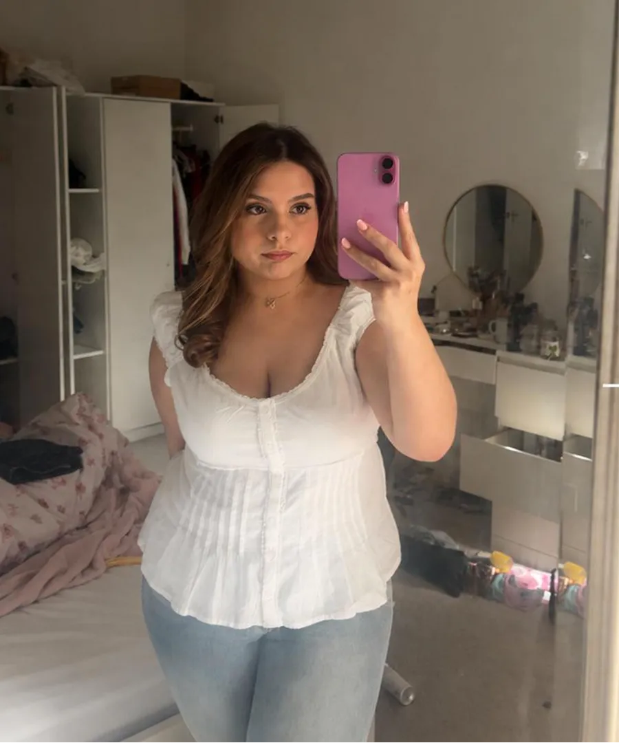 Woman taking a mirror selfie in a bedroom, wearing a white sleeveless blouse and light blue jeans, holding a pink phone with her right hand.