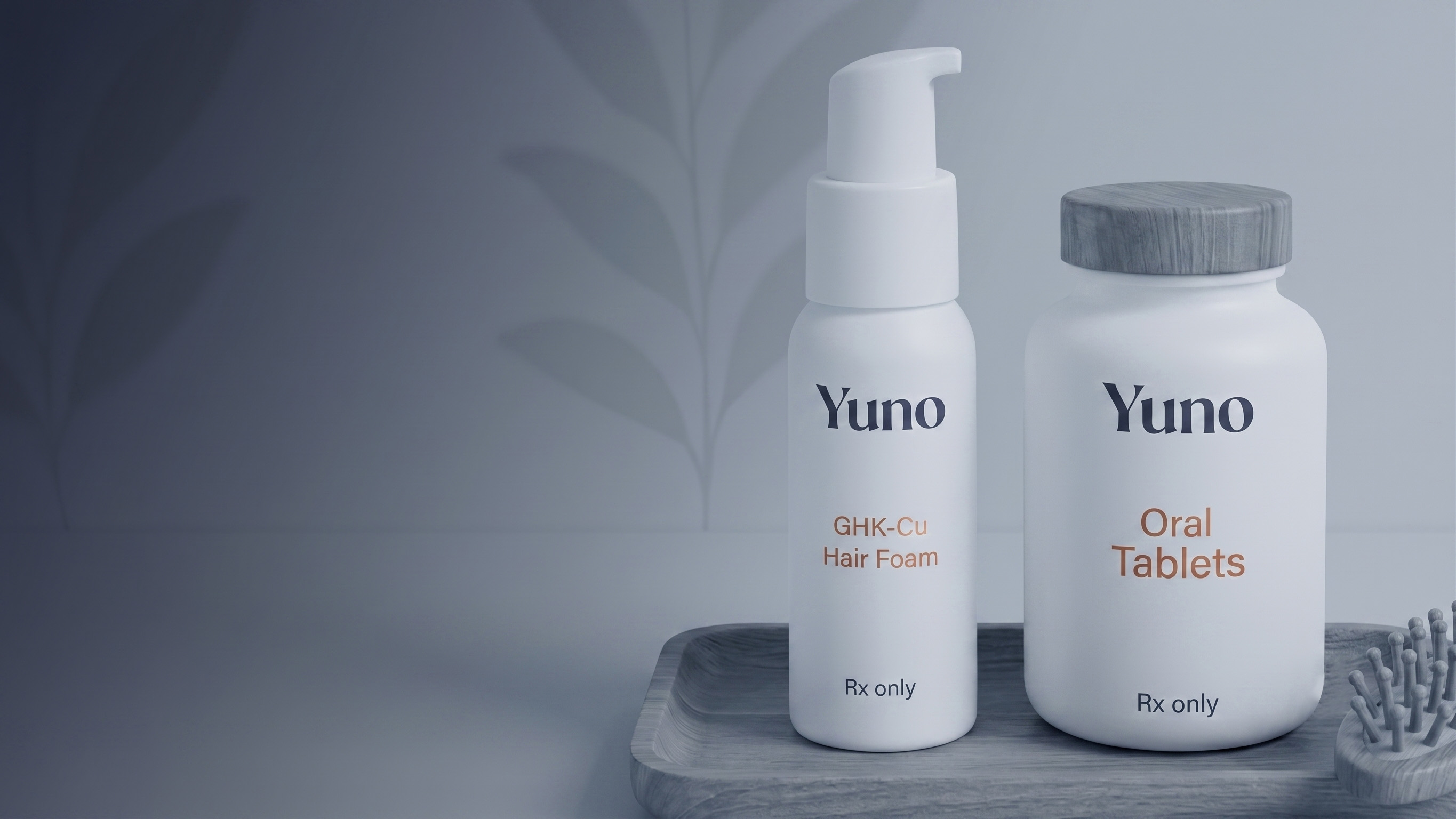 Two white Yuno products on a gray tray: a pump bottle labeled GHK-Cu Hair Foam and a jar labeled Oral Tablets, both marked Rx only, with a hairbrush nearby and a subtle leaf pattern on the background wall.