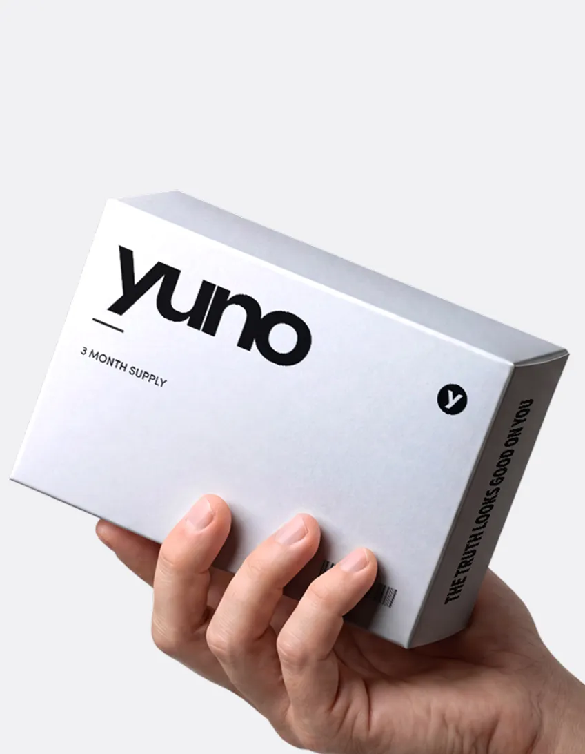 Hand holding a white box labeled 'yuno' with text indicating it is a 3 month supply, with a small circular logo containing 'y' and side text reading 'THE TRUTH LOOKS GOOD ON YOU'.