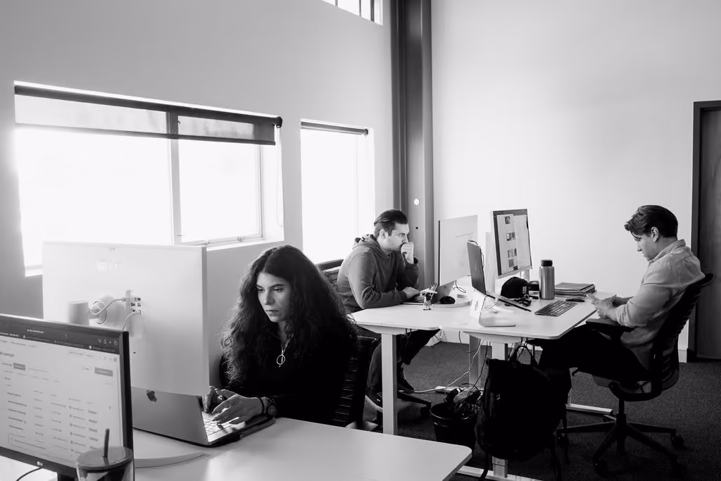 Three people working on laptops and computers in a bright office with large windows.