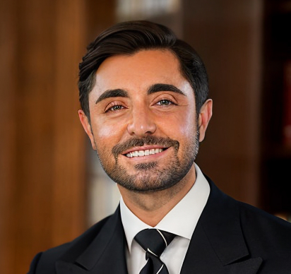 Smiling man with dark hair and beard wearing a suit and striped tie against a blurred background.