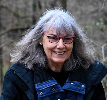 Smiling older woman with gray hair and glasses wearing a blue jacket outdoors in a forest.