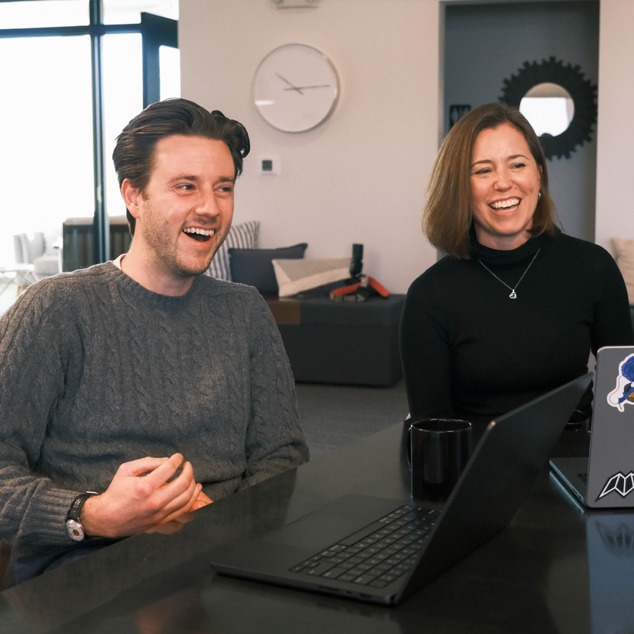 Man and woman laughing together while sitting at a table with laptops in a bright office.