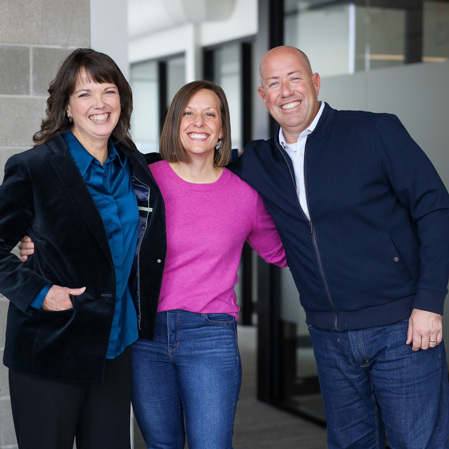 Three people smiling and posing together indoors, two women and one man wearing casual and business attire.