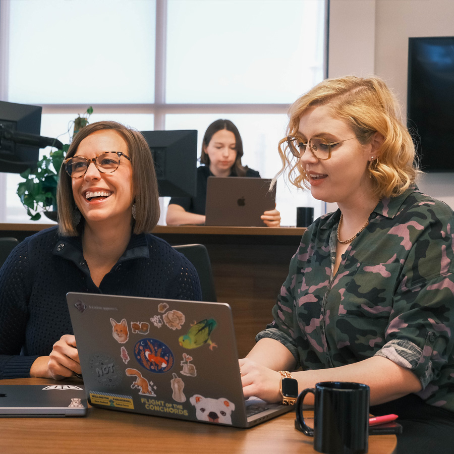 Two women with glasses working on laptops in an office while a third person works in the background.