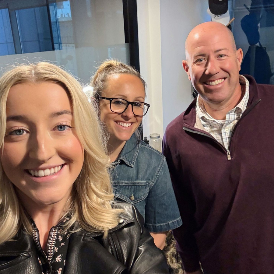 Three smiling adults pose together indoors, one wearing glasses and another with a leather jacket.