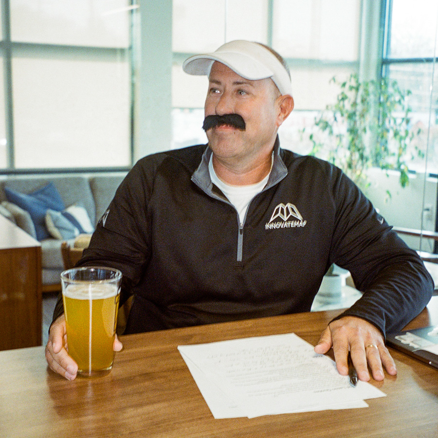 Man in white visor and black jacket with mustache holds beer glass sitting at a table with papers.