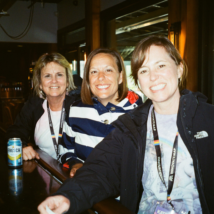 Three smiling women wearing lanyards sitting together at a bar with a can of Fresca soda nearby.