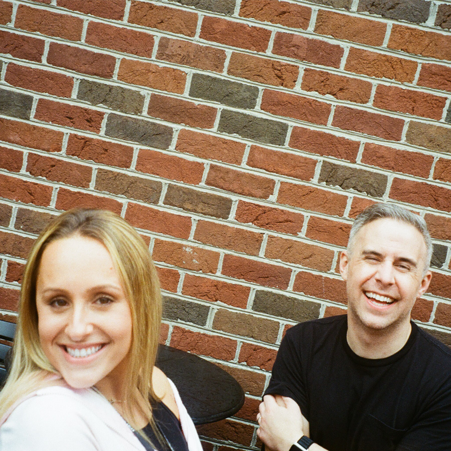 Smiling man and woman sitting outdoors against a red and gray brick wall.