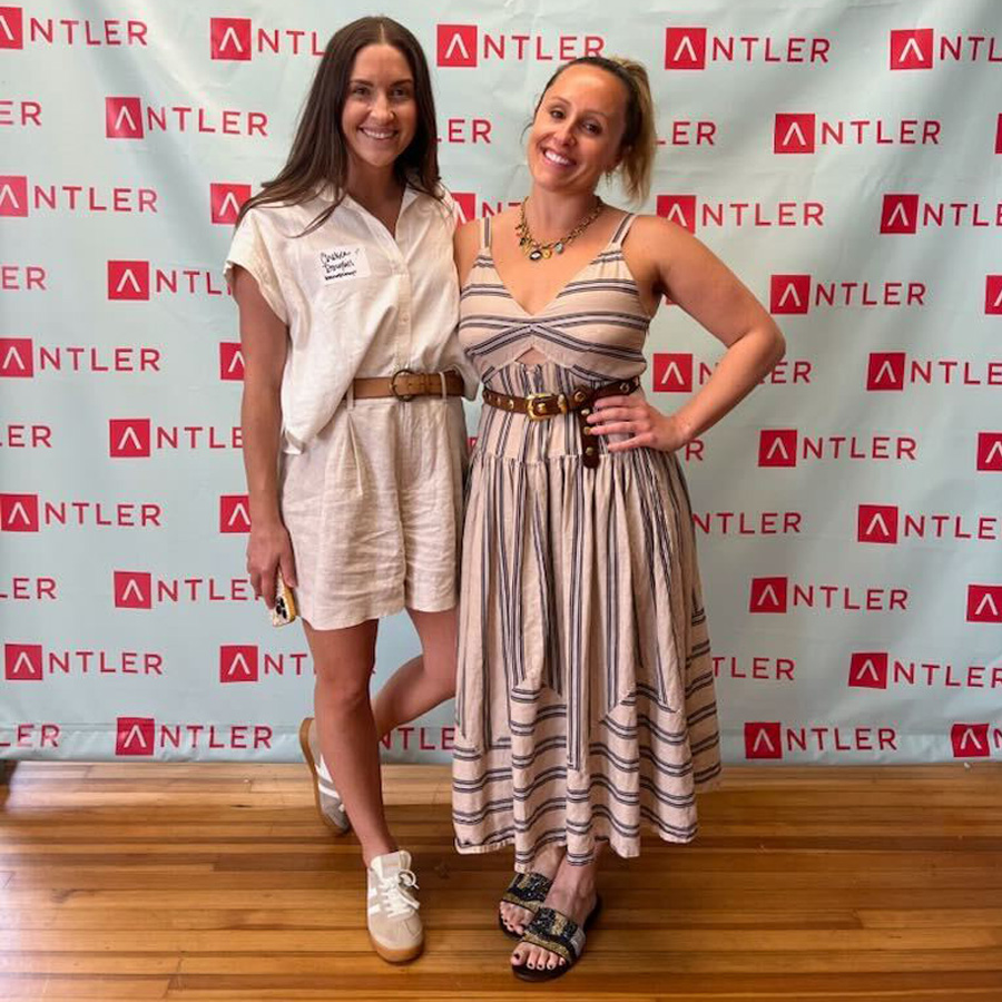 Two smiling women stand side by side on wooden floor with an Antler branded backdrop behind.