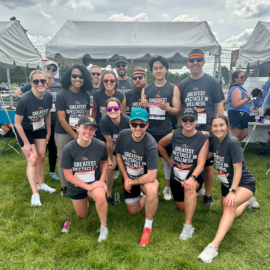 Group of runners wearing matching wellness event shirts posing on grass with tents in background.