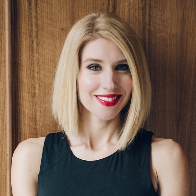 Smiling woman with straight blonde hair wearing a black sleeveless top against a wooden background.
