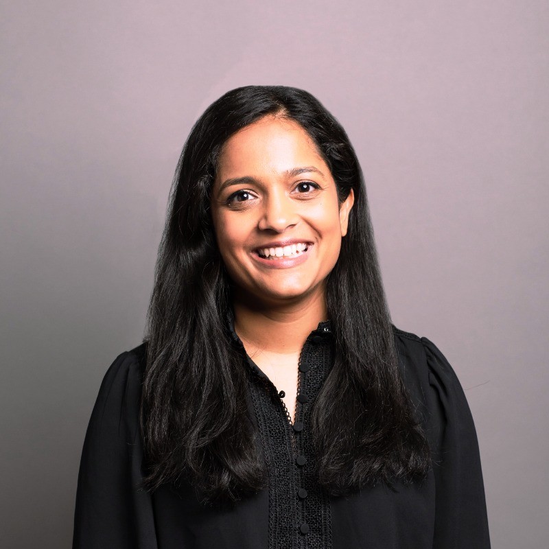 Smiling woman with long black hair wearing a black buttoned blouse against a gray background.
