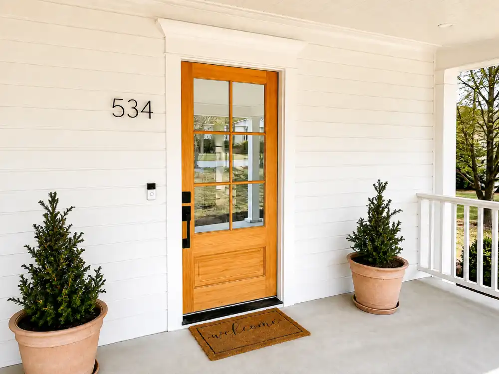 Wood front door with glass panes on a white covered porch with potted evergreens