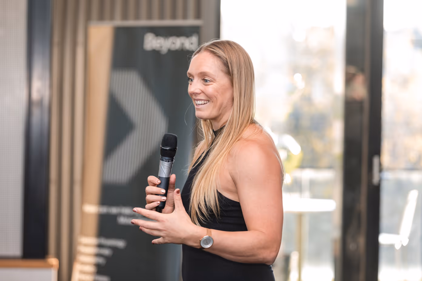 Woman with long blonde hair speaking and holding a microphone at an indoor event.