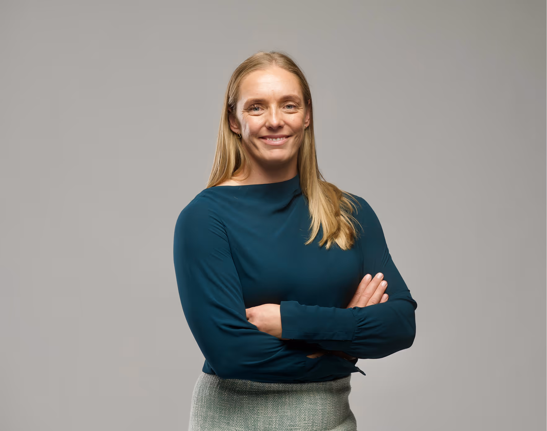 Smiling woman with long blonde hair wearing a dark teal top and gray skirt with arms crossed.