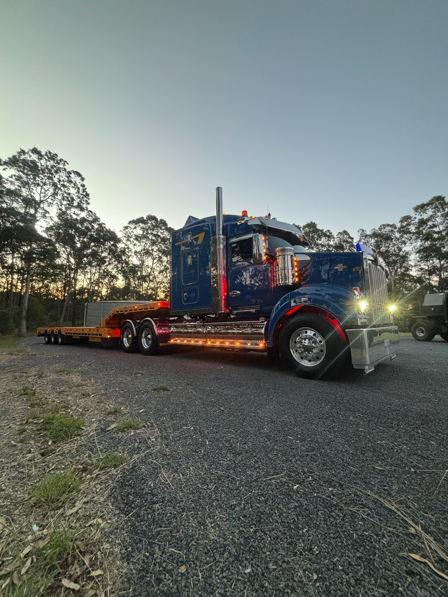 Blue semi-truck with trailer parked on gravel road at dusk with trees in background.