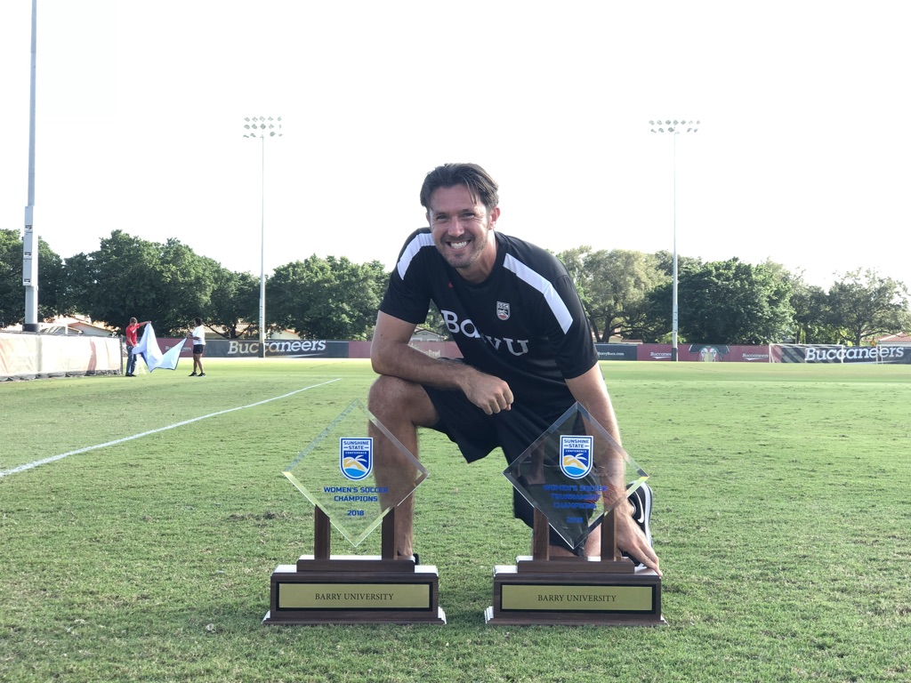 Brian Galvin posing with two soccer championship trophies on a soccer field