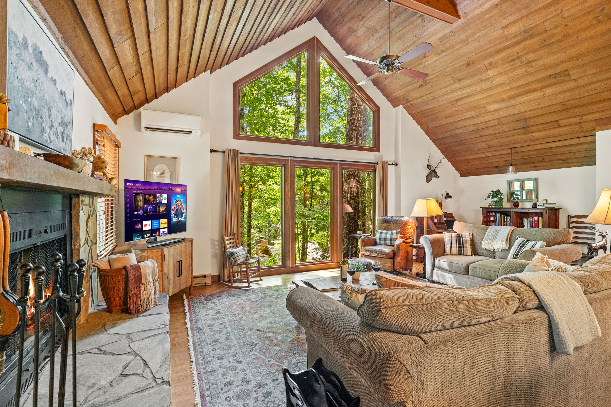 The great room with a wood-burning fireplace, a ceiling fan under the vaulted ceiling, and tall windows onto the woods