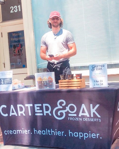 Man standing behind a Carter & Oak Frozen Desserts table with product samples and signage outdoors.