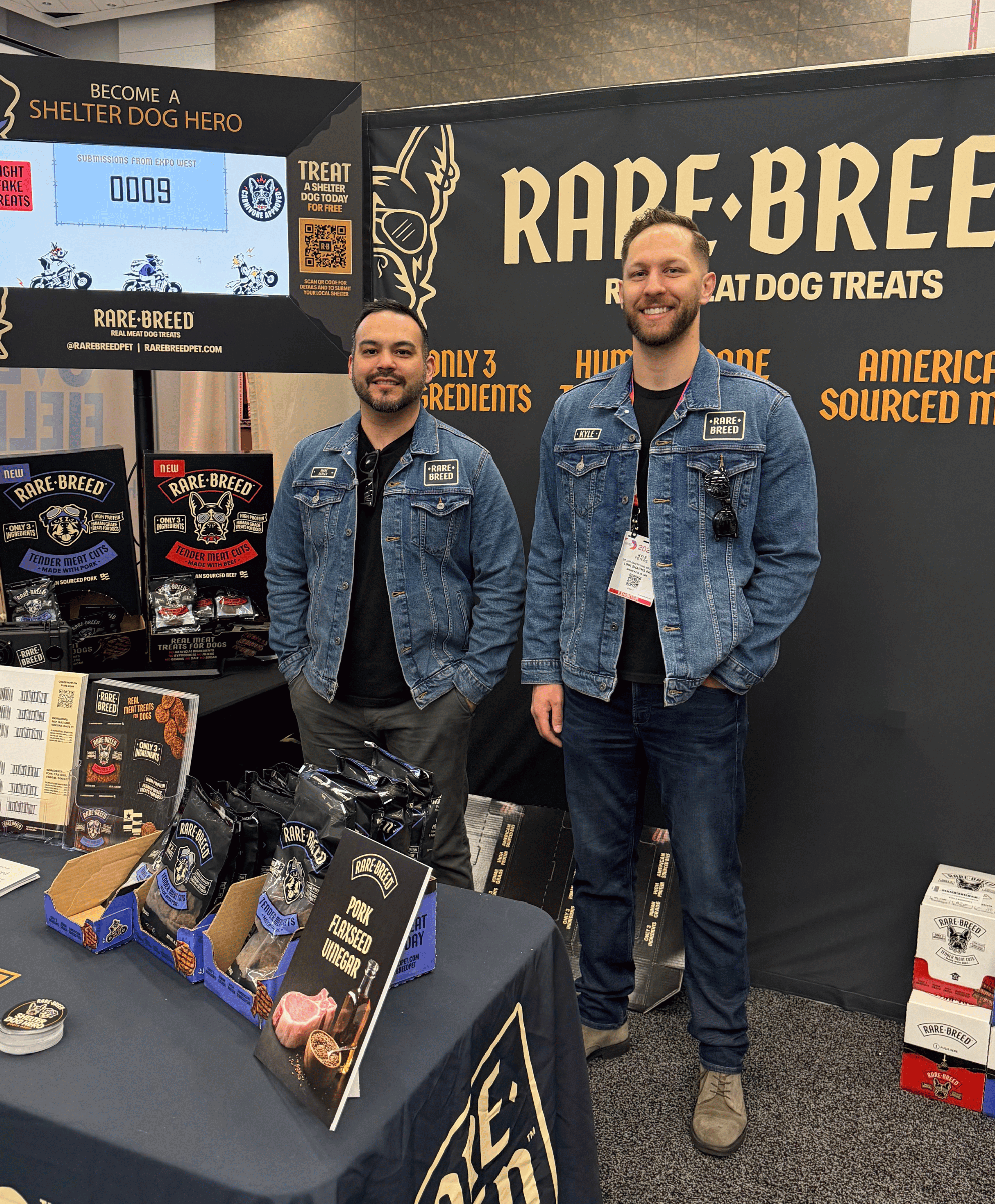 Two men in denim jackets stand at a Rare Breed dog treats booth with product displays and signage.