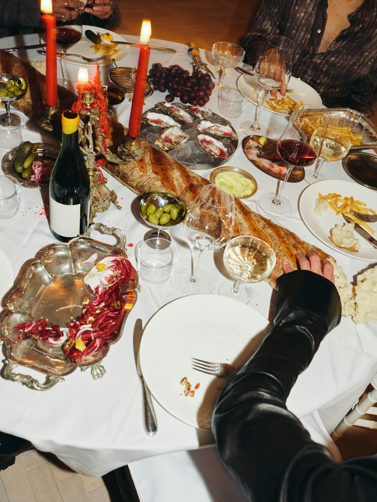 Table with long baguette, oysters, grapes, olives, red wine, fries, candles, and people seated at dinner.