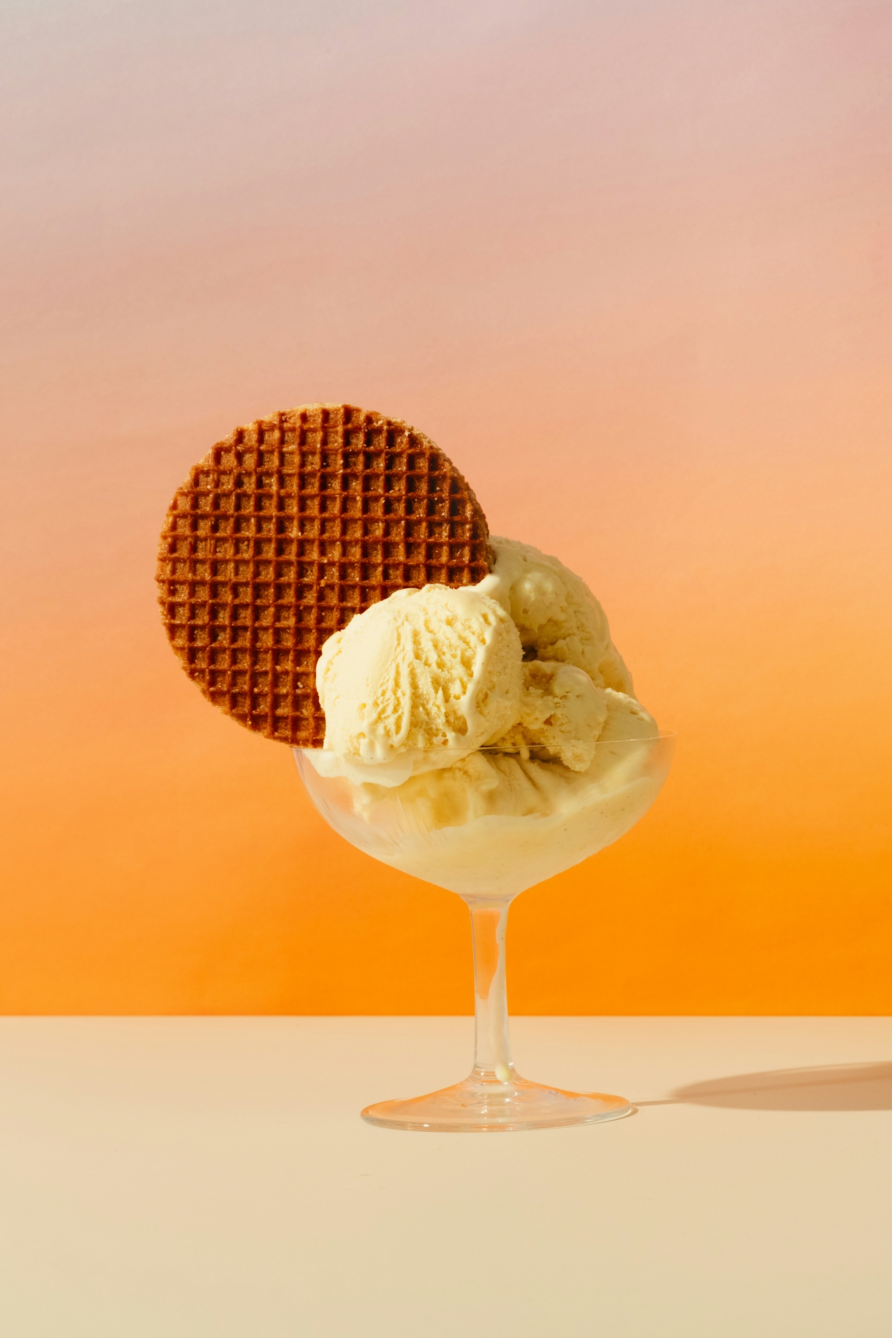 Vanilla ice cream scoops in a glass dish with a round stroopwafel against an orange gradient background.