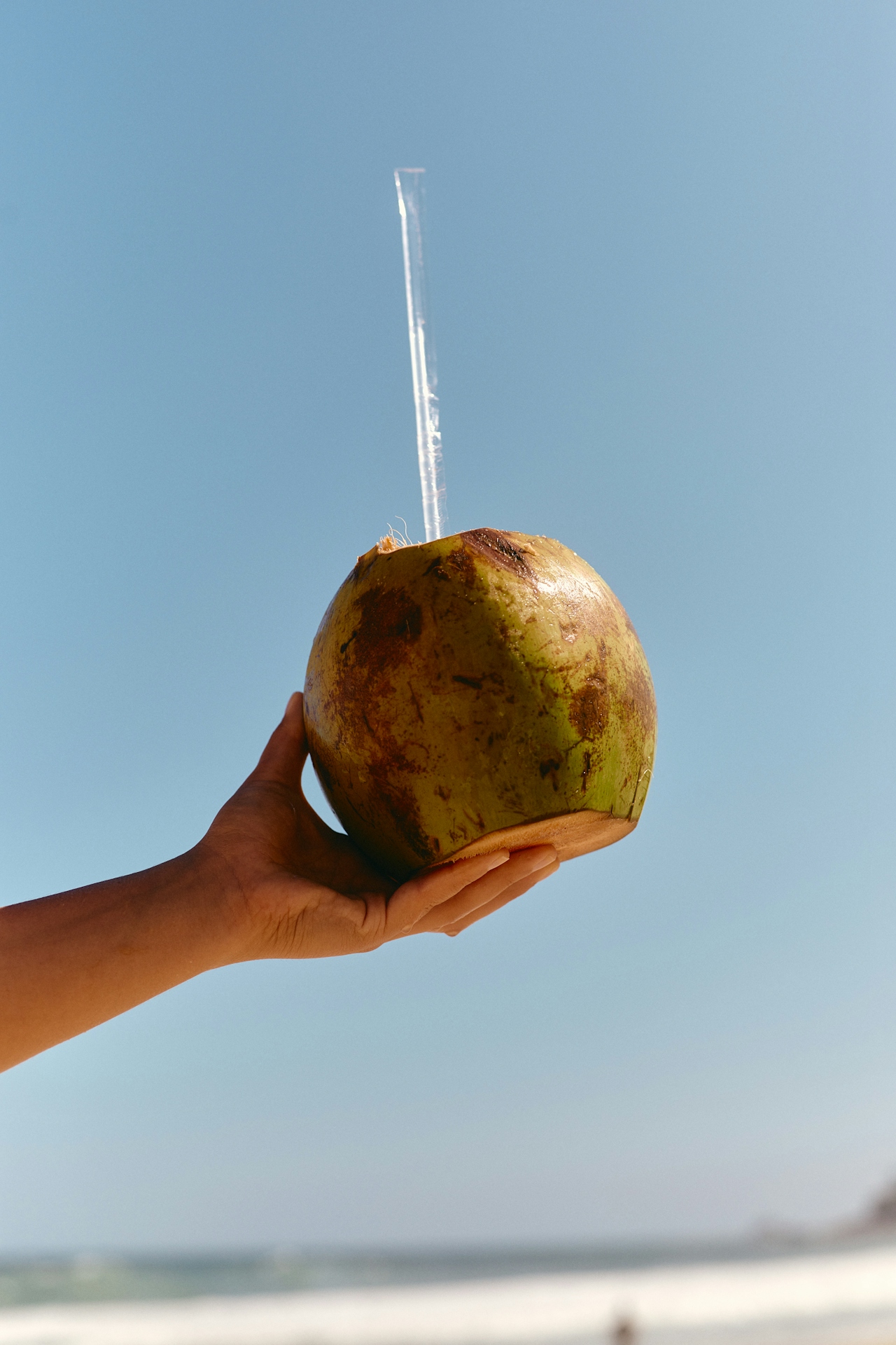 Hand holding a green coconut with a straw against a clear blue sky at the beach.