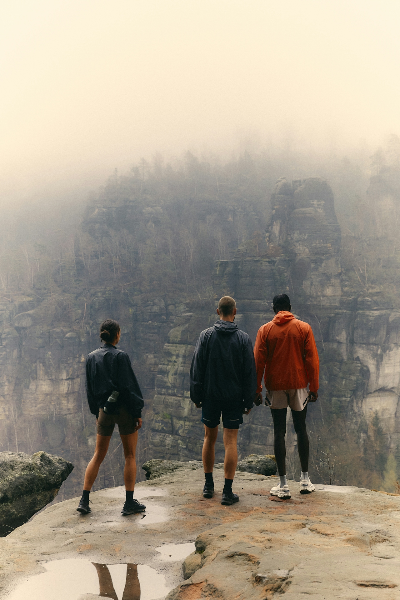 Three hikers standing on rocky cliff edge looking at fog-covered forest and rock formations.