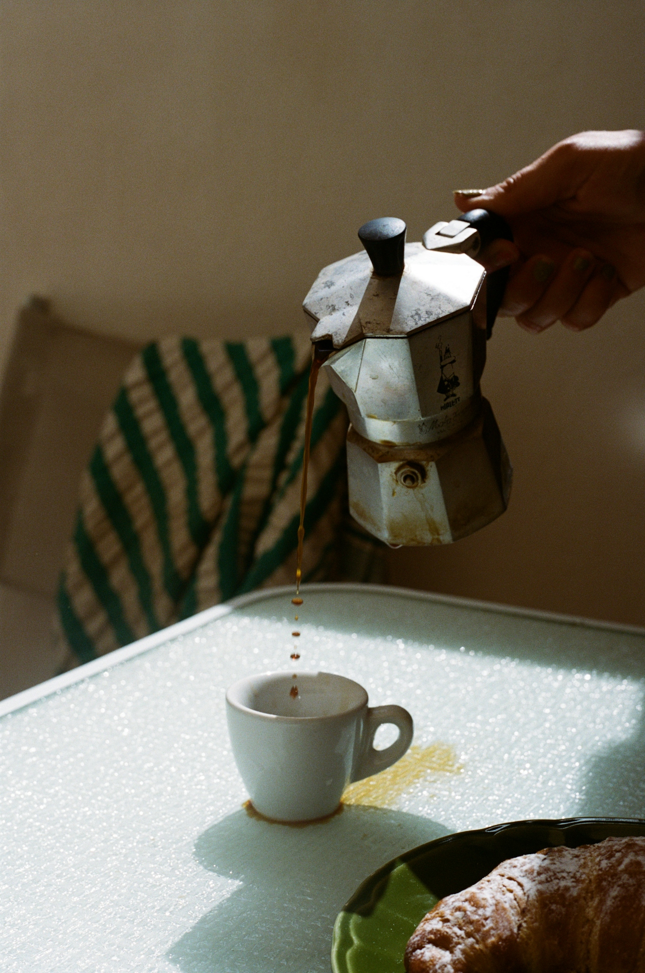 Coffee pouring from a moka pot into a small white cup on a glass table with a croissant nearby.