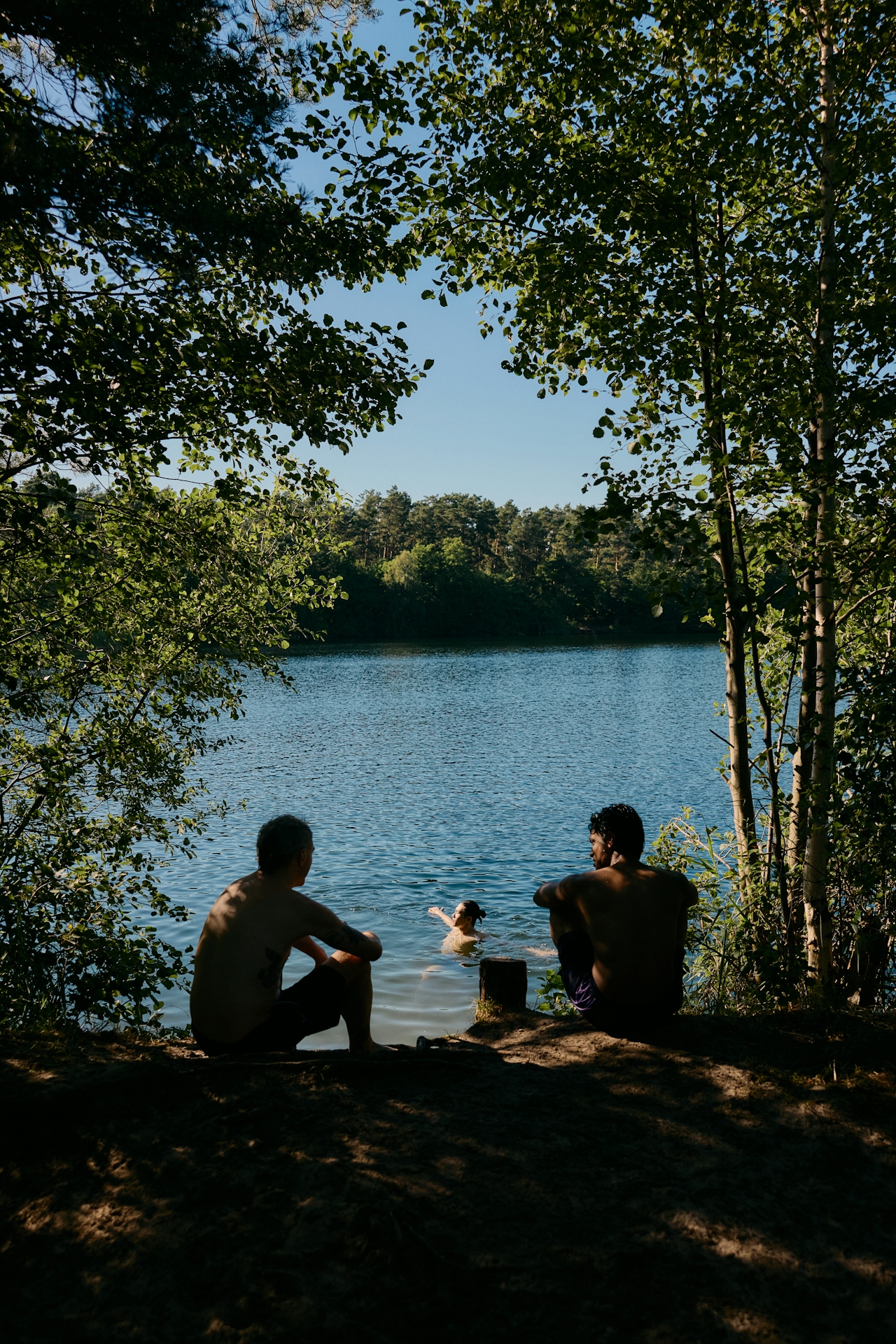 Two shirtless men sitting by a lake watching a woman swim surrounded by trees on a sunny day.