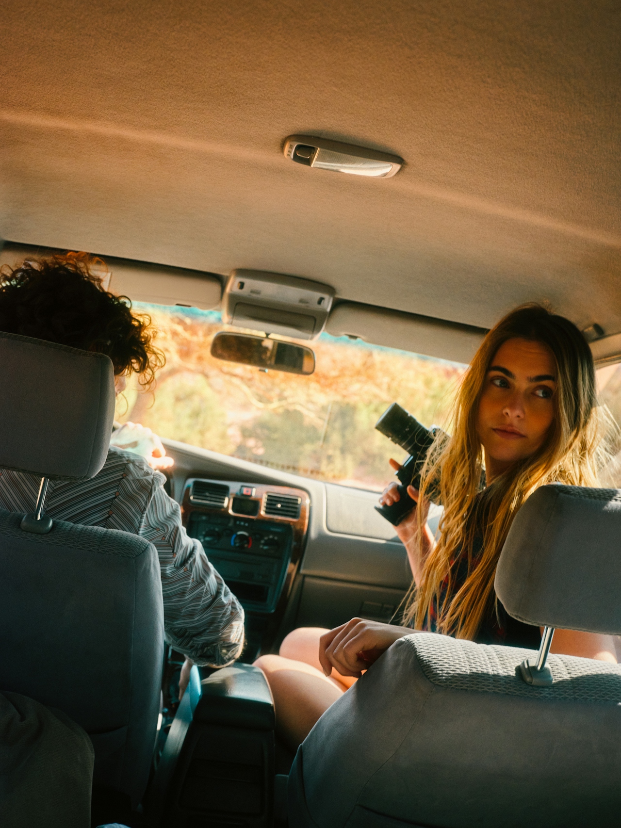 Woman with long hair holding a camera inside a car while a person drives ahead.