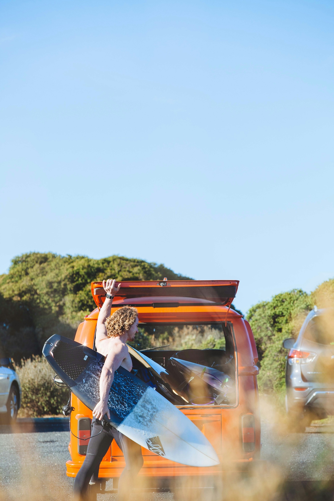 Man holding surfboard standing by open back of orange van in sunny outdoor parking area.