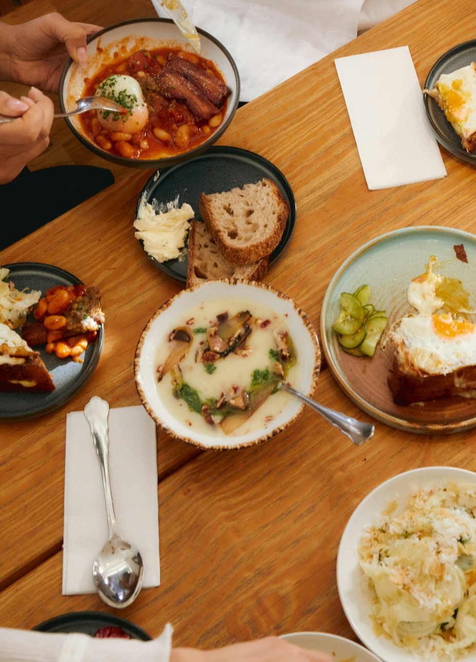 Wooden table with plates of eggs, baked beans, bread, mushrooms, and salad, hands visible eating.