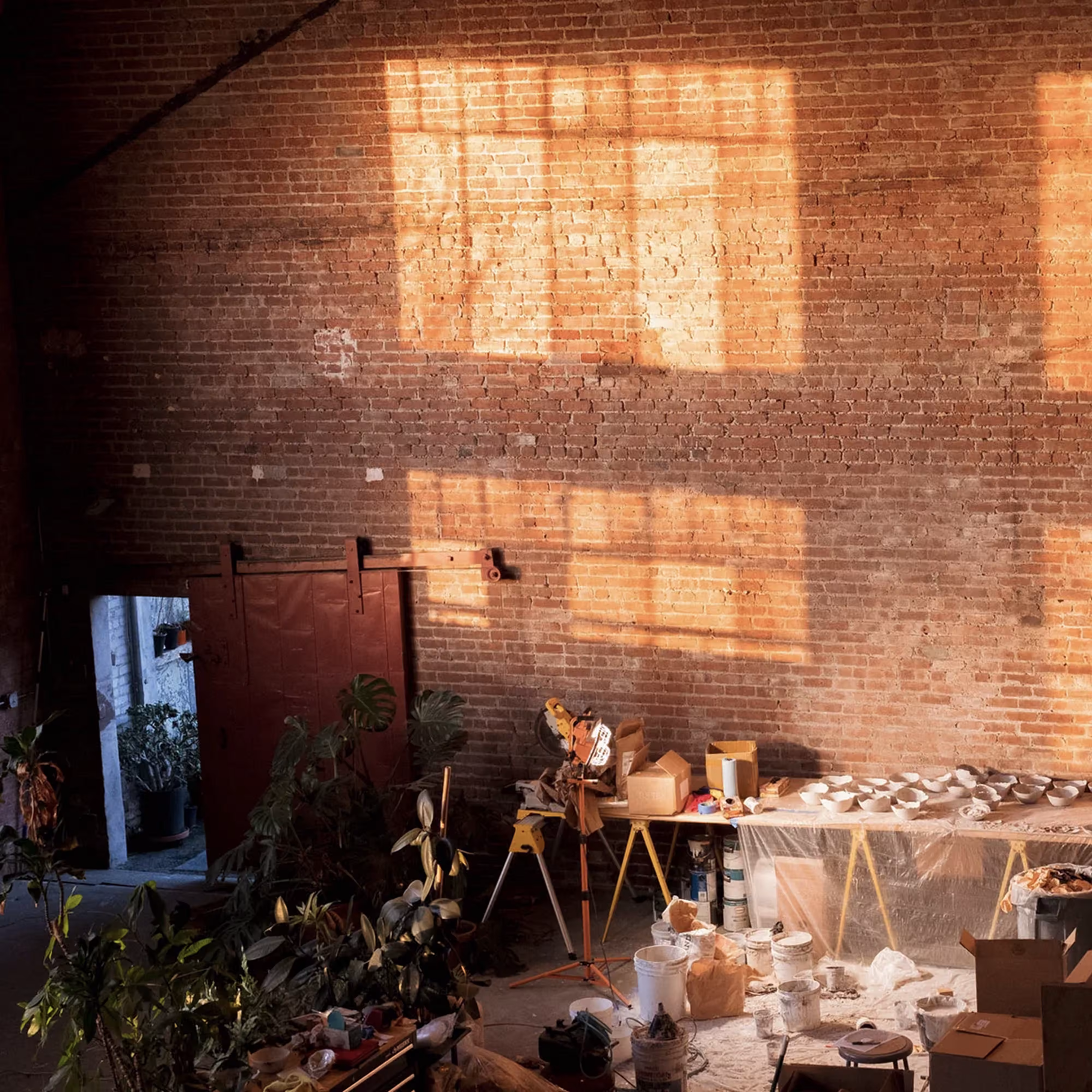 Sunlight casts window shadows on a brick wall in a cluttered workshop with plants and tools.