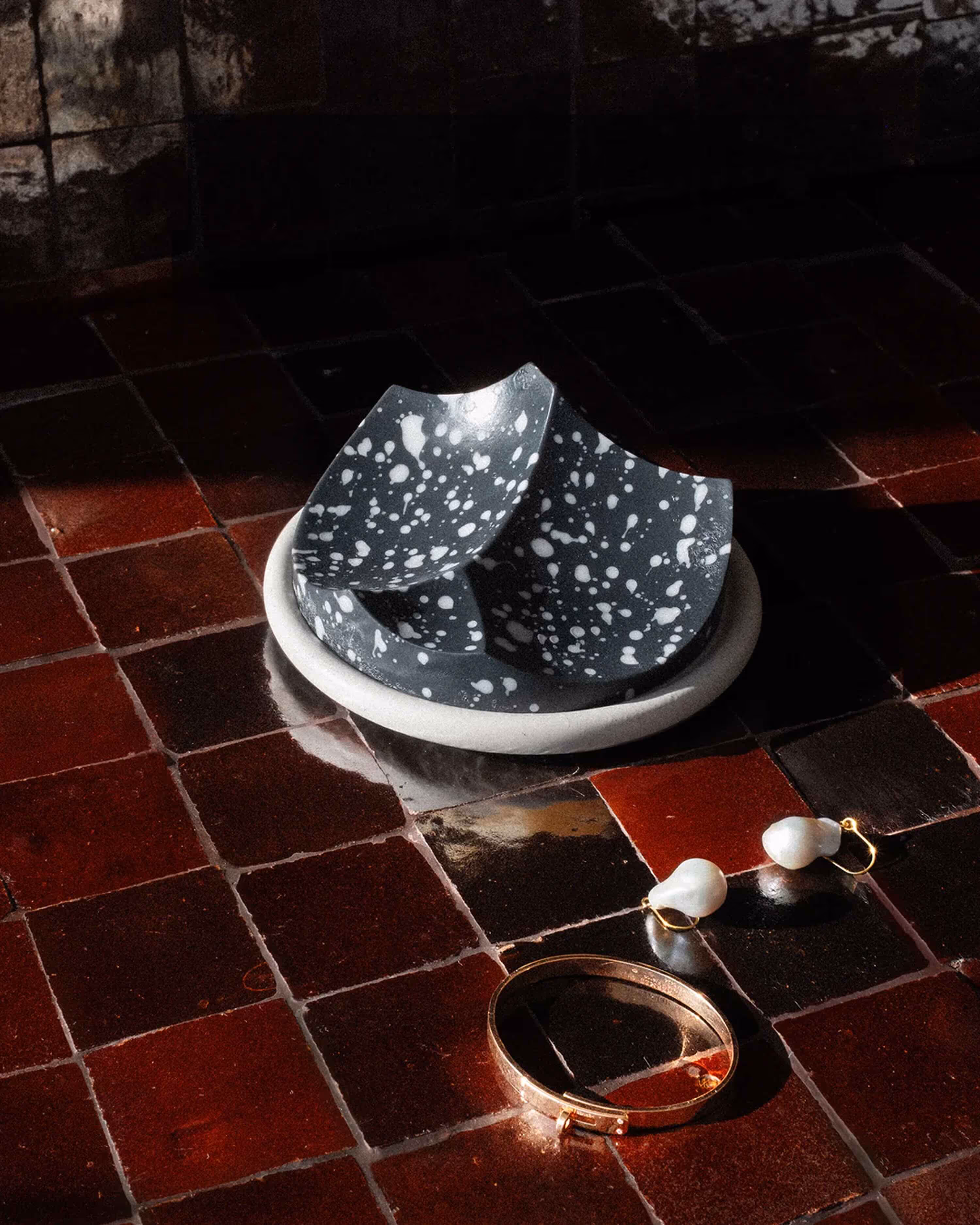 Speckled black and white ceramic dish on red tiled surface with pearl earrings and gold bracelet.