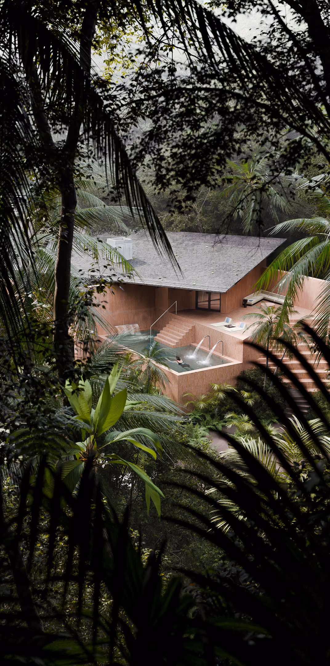 A pink sand-colored building with outdoor Thalassotherapy pool is seen through lush tropical forest vegetation.