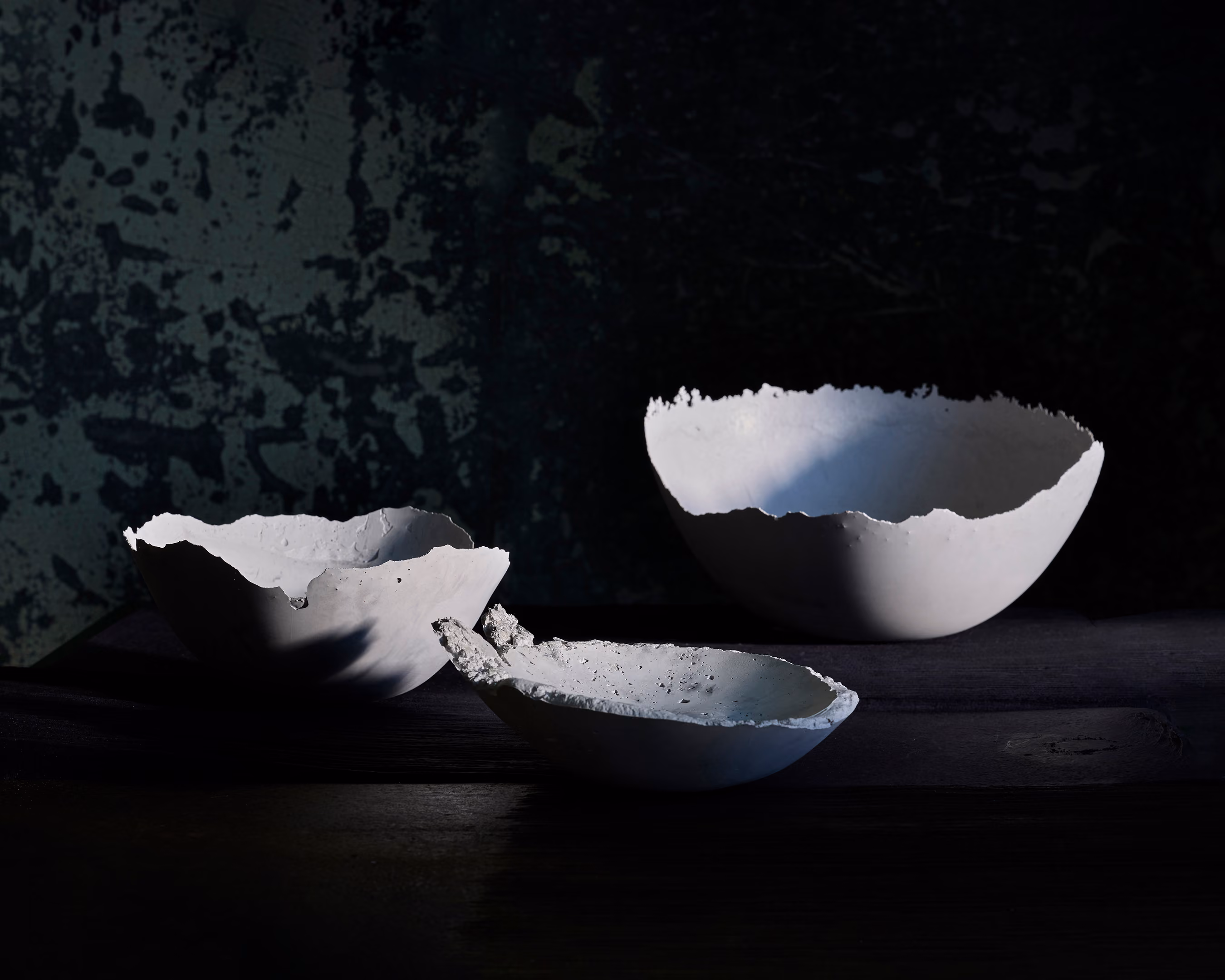 Three delicate, rough-edged, white concrete bowls sit on a dark surface against a dark background.