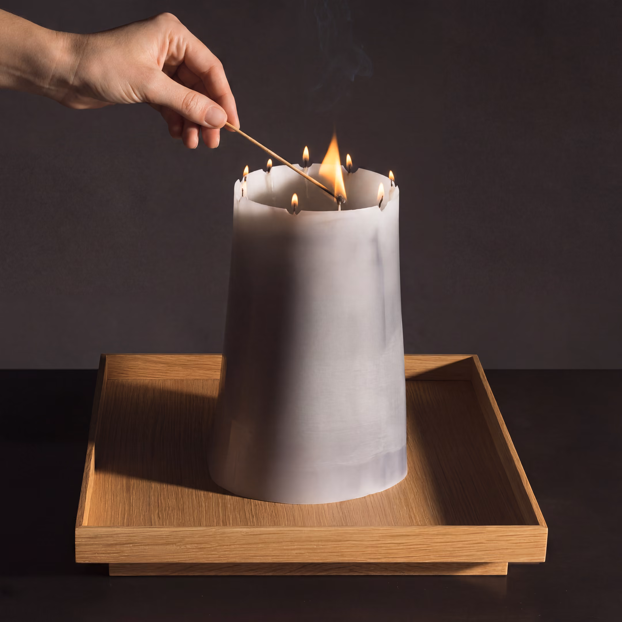 Large candle with multiple wicks on top is lit with a long match. The candle sits on a square wood tray against a dark background.