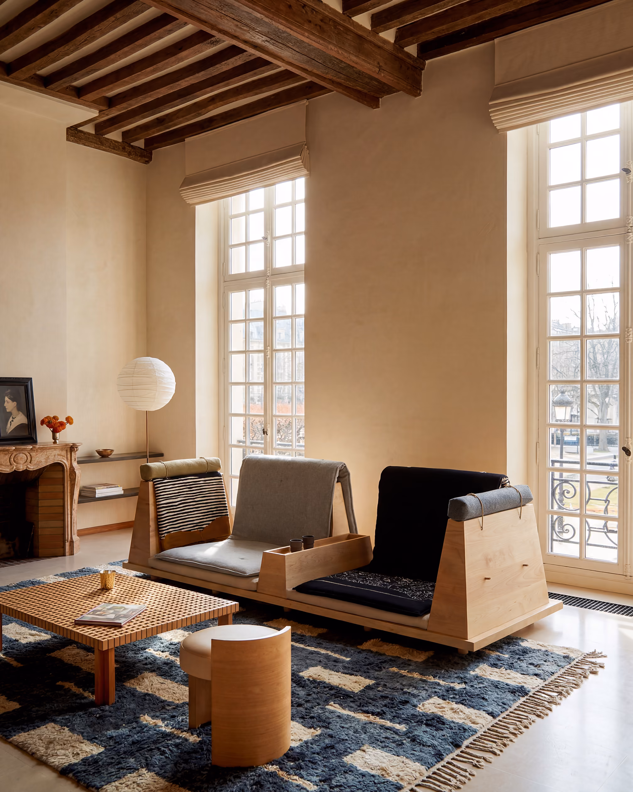 Warm, modern living room with wood Zabuton sofa, patterned rug, coffee table, large windows, and wood ceiling beams.