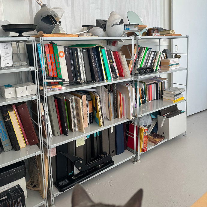 Metal shelves filled with books, binders, files, and concrete objects in a studio setting.