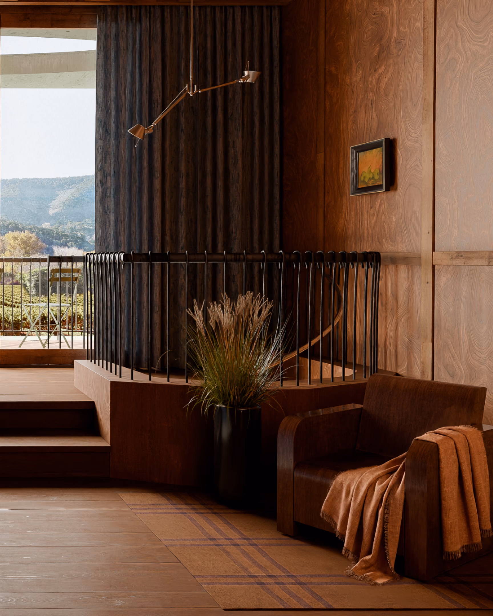 Modern cozy room with brown armchair, blanket, potted grass, and balcony overlooking green hills.
