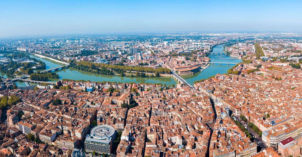Vue aérienne de Toulouse avec la Garonne traversant la ville et des toits rouges sous un ciel bleu clair.