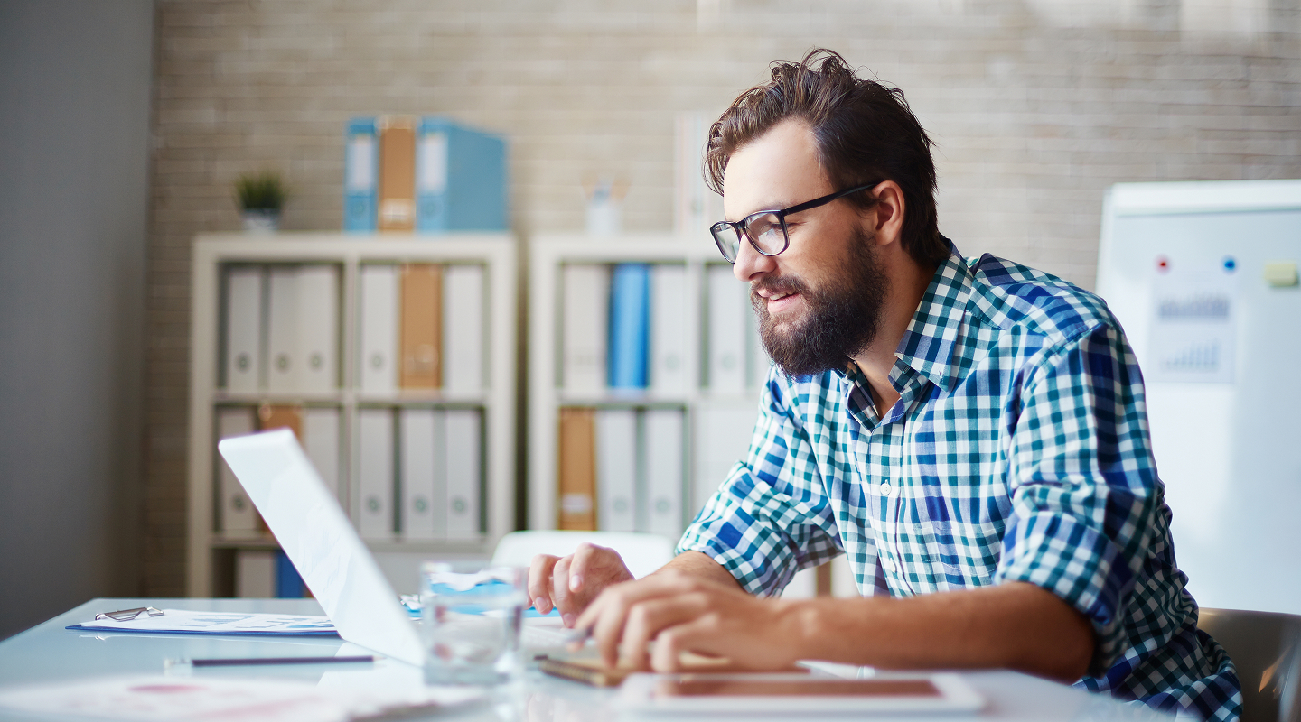Bearded man in glasses and checkered shirt working on a laptop in an office with shelves and a whiteboard.